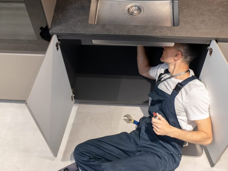 Solution. Top view of man plumber in overalls holding ball valve in hand lying on floor near open set of tools in kitchen fixing breakdown of sink